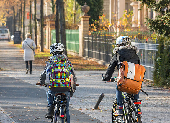 Zwei Kinder mit Rucksäcken fahren auf einem baulich getrennten Radweg in einem Wohngebiet