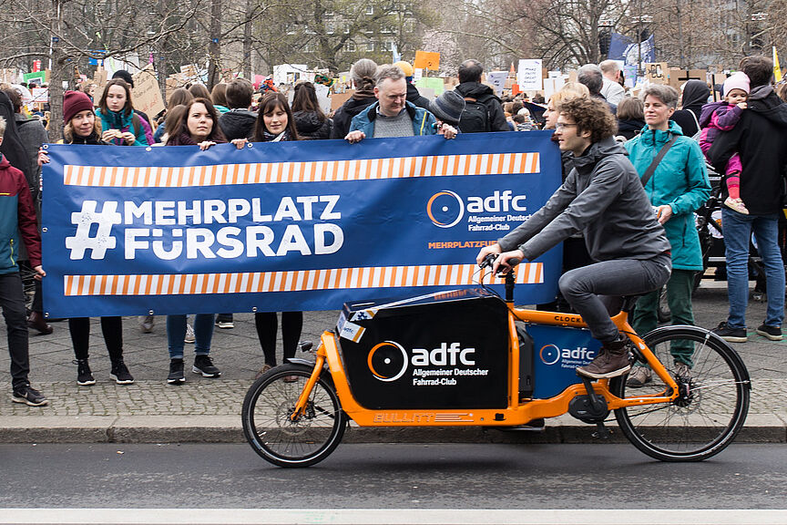 Ein Mensch auf einem Lastenrad vor einer Gruppe, die ein "Mehr Platz fürs Rad"-Banner hoch hält.