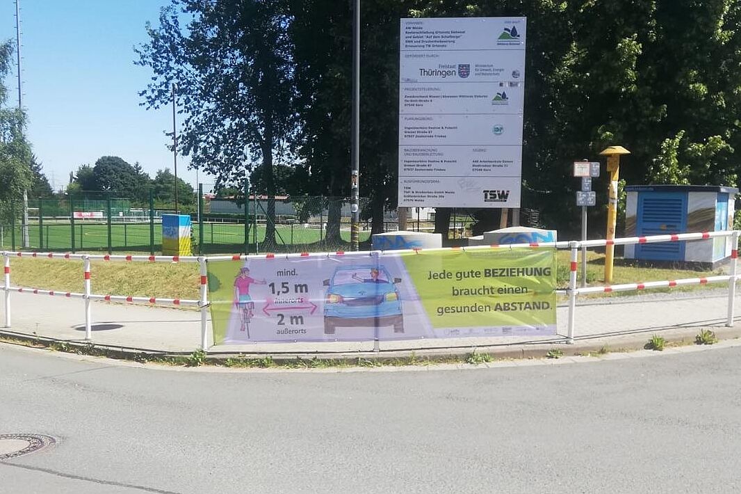 Ein Abstandsbanner hängt an einem Zaun an der Straße.
