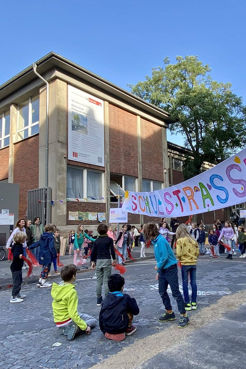 Schulstraße Kinder verschiedenen Alters spielen und bewegen sich auf einer gepflasterten Straße vor einem zweistöckigen Backsteingebäude. Über der Straße hängt ein buntes Banner mit der Aufschrift „Schulstraße“ und einem grünen Baum-Symbol. Im Hintergrund sind Erwachsene zu sehen, die die Aktion begleiten.