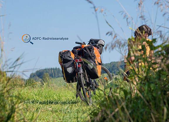 ADFC-Radreiseanalyse 2023 Reiserad mit Packtaschen steht in einer gemähten Wiese, Frau hinter hochgewachsenem Gras